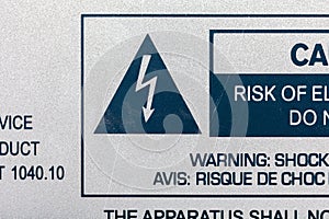 A close-up view of a bilingual warning label detailing the risk of electrical shock from a machine device or appliance, with a