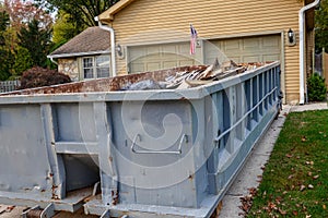 Closeup view of a blue dumpster filled with construction debris in the driveway of a yellow house in front of the garage doors