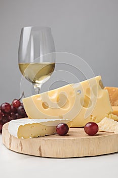 Close up view of brie and maasdam cheese on wooden board, grapes and wine glass on gray