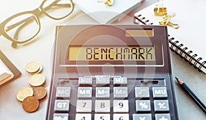 Close-Up View of a Calculator Displaying the Word BENCHMARK on a Desk Surrounded by Financial Paraphernalia