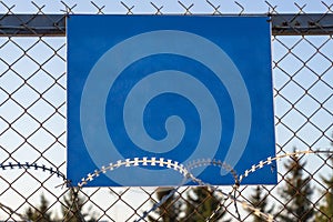Close-up view of a caution banner, mock-up of a blue plate on an iron mesh fence with barbed wire. Closed area designation blank.