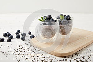 close up view of chia puddings with fresh blueberries and mint on wooden cutting board on white tabletop