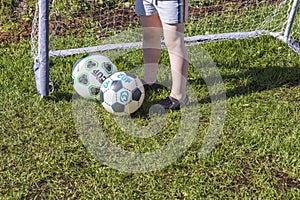 Close up view of the child`s feet near two balls on football goal background.