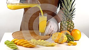 The close-up view of the cooker pouring the orange juice at the high speed into the glass placed near fruit tropical