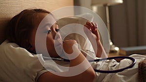 Close-up view of cute little girl playing with stethoscope while lying in bed in bedroom.
