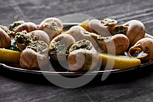 Close up view of delicious cooked escargots with lemon slices on plate on black wooden table.