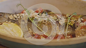 Close up view of fresh baked fish lying in deep plate decorated with lemon, tomatoes cherry, greens and nuts. Food