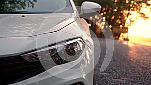 Close up view of front light of car headlights. unrecognizable white car is parked on side of road with setting sun in