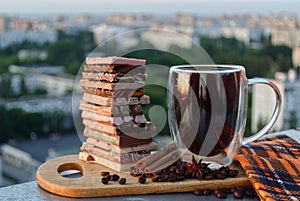 Close-up view of a glass mug with black coffee and a stack of pieces of various chocolate, coffee beans, cinnamon sticks
