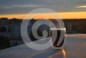 A close-up view of a glass mug with hot black coffee standing on a steel parapet against a blurred background of dawn sky