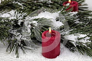 Close up view of a glowing red candle with snow covered fir tree branches for a merry Christmas or happy New Year holiday