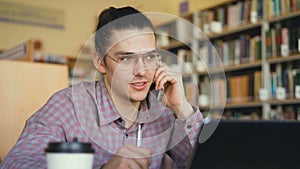 Close-up view of handsome young hipster male student wearing glasses sitting in university library talking on smartphone