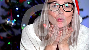 Close-up view  of happy woman in red santa hat sending air kiss over christmas tree lights background. Woman looking camera and wa