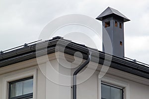 Close-up view of house with gray fold roof and plums and filing of roof overhangs with soffits and brick stone pipe covered with