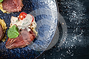 Close up view on Juicy medium roast beef steak sliced with blood served on dark plate with tomato and mashed potato. Meat food.