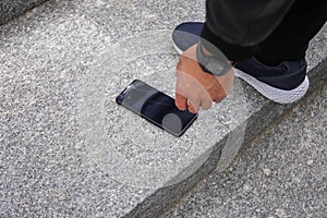 Close up view of kneeling man in jeans and shoes picking up broken phone on paved sidewalk outdoors