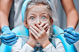 Close up view of a little girl looking scared and terrified screaming covering her mouth from the dentists with medical tools