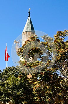 Close-up view minaret which is covered by green tree leaves. Blue sky background. Turkish flag near the minaret