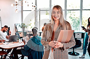 Close up view portrait of pretty cheerful business woman in an office environment holding laptop. People working background