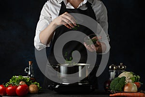 Close-up view of the professional chef in black apron adding chopped parsley in boiling water for soup on dark blue background.