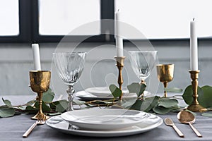 close up view of rustic table setting with wine glasses, eucalyptus, vintage cutlery, candles in candle holders and empty plates