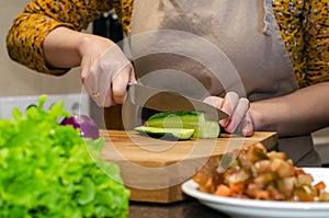 Close-up view of the salad preparation process. A woman in an apron cuts a cucumber on a wooden cutting board