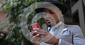 Close up view of serious bussinessman in hat and suit typing message and looking at phone screen. Serious guy in stylish