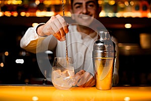 close-up view on shaker and bartender stirring alcoholic cocktail with bar spoon on yellow bar