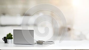 Close up view of simple workspace with laptop, notebooks, coffee cup and tree pot on white table with blurred office room