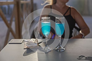 Close-up view of two glasses with blue alcoholic beverages and a girl sitting at a table in a restaurant.