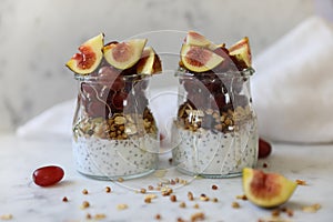 Close up view of two glasses filled with yogurt with chia seeds, granola, fresh figs and purple grapes drizzled with honey against