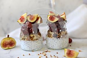 Close up view of two glasses filled with yogurt with chia seeds, granola, fresh figs and purple grapes drizzled with honey against