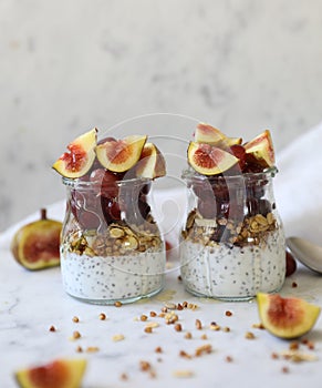 Close up view of two glasses filled with yogurt with chia seeds, granola, fresh figs and purple grapes drizzled with honey against