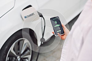 Close up view. Woman charging her electric car and monitoring process on smartphone
