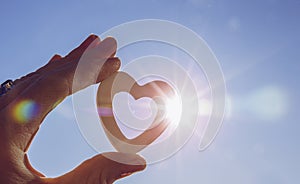 Close up view of woman hand holding white wooden heart against sun and blue sky with lens flare and sun`s photography star effect.