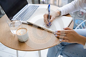 Close-up view of a woman`s hands writing notes in a notebook. Girl working in a cafe.