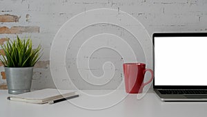 Close up view of workplace with blank screen laptop, stationery, red mug and tree pot on white table