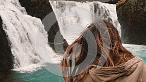 Close-up view of young attractive woman standing alone and looking on powerful waterfall in Iceland, hair wave on wind.