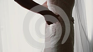 Close-up view of young bride puts a bow on her wedding dress. Woman standing in front of window and puts on the suit.