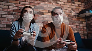 Close-up view of young couple in medical face masks playing video games on console sitting together.