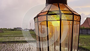 Close-up of a vintage lamp with flickering light on background of rice fields at sunset with pink sky. Antique lamp with