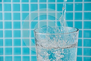 Close-up of water falling into a glass, with unfocused light blue mosaic background, horizontally
