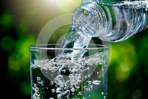 Close up of water flowing from drinking water bottle into glass on blurred green nature bokeh background with soft sunlight