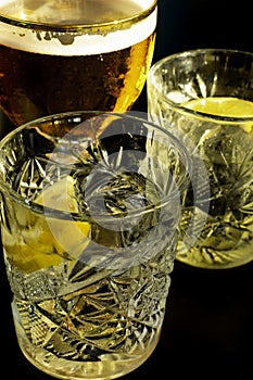 Close up of a wet glass of cold light beer with foam and vermouth Martini alcohol cocktail with yellow lemon slice and ice cubes o