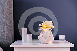 Close up white bedside table with ceramic female body-shaped vase with rose flower and candles on dark blue wall