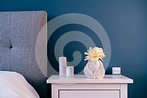 Close up white bedside table with ceramic female body-shaped vase with rose flower and candles on dark blue wall