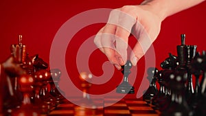 Close up of white and black wooden chess pieces on board. Man's hand makes move of black pawn on chessboard on a red