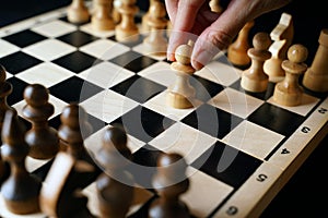 Close up of white and black wooden chess pieces on board. Woman's hand makes first move of white pawn on chessboard