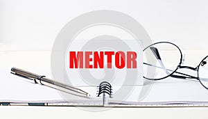 Close-up of a white card with the text MENTOR, a stylish pen and glasses on a desktop