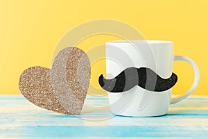Close up of white ceramic cup with a felt moustache and sparcle golden heart on yellow background. Copy space
